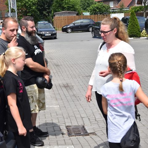 Vierte Personen stehen auf einem Parkplatz, zwei Kinder in Sportkleidung, Erwachsene sprechen miteinander.