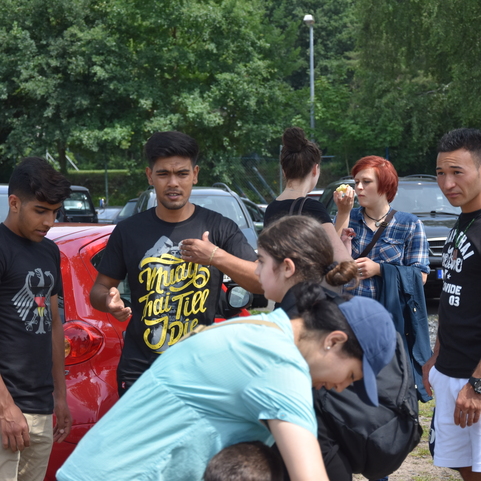 Gruppe von f&uuml;nf Personen, die an einem Parkplatz stehen, umgeben von Autos und B&auml;umen.