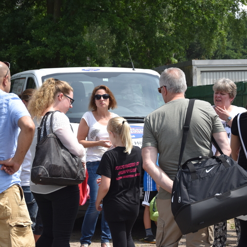 Gruppengespr&auml;ch von mehreren Personen im Freien, umgeben von Fahrzeugen und B&auml;umen, bei sonnigem Wetter.