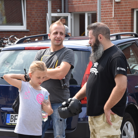 Drei Personen stehen vor einem Auto: ein Mann mit Bart, ein Mann mit T-Shirt und ein M&auml;dchen mit Wasserflasche.