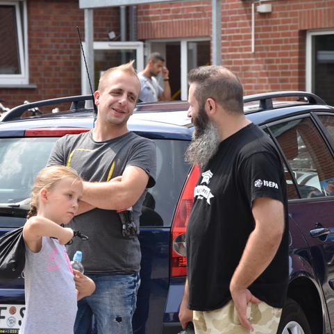Drei Personen stehen vor einem Auto, zwei M&auml;nner unterhalten sich, w&auml;hrend ein M&auml;dchen aufmerksam zuschaut.