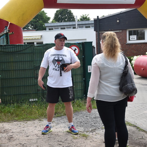 Eine Gruppe von Menschen steht unter einem bunten Tor, das zu einer Veranstaltung f&uuml;hrt. Ein Mann spricht mit zwei Kindern.