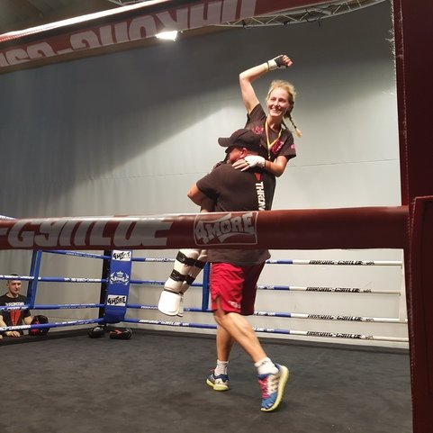 Siegerin feiert ihren Triumph in einem Boxring, w&auml;hrend ihr Trainer sie hochhebt und Zuschauer im Hintergrund zuschauen.