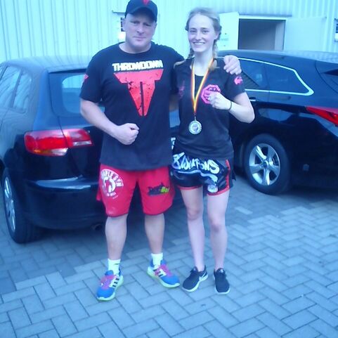 Trainer und Athletin stehen l&auml;chelnd vor einem schwarzen Auto, die Athletin tr&auml;gt eine Medaille und Sportkleidung.