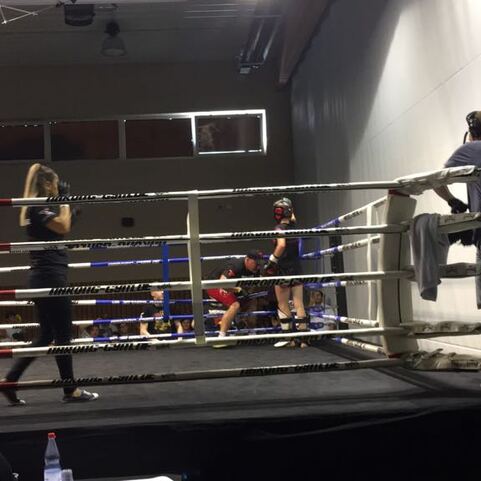 Boxring mit K&auml;mpfern und Trainern; eine Frau betritt den Ring, ein Trainer beobachtet vom Rand aus.