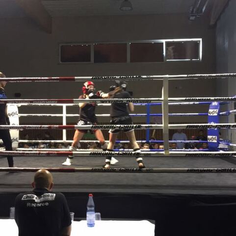 Zwei Boxer k&auml;mpfen im Ring, umgeben von Zuschauern und Kameras, w&auml;hrend eine Schiedsrichterin beobachtet.