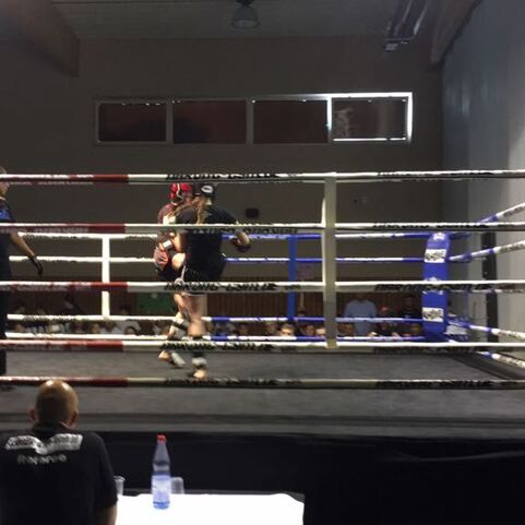 Kampfszene im Ring mit zwei K&auml;mpfern, einer davon wird von einer Trainerin unterst&uuml;tzt, w&auml;hrend ein Kameramann filmt.