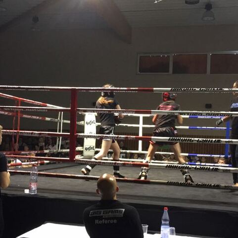 Zwei Personen im Boxring k&auml;mpfen gegeneinander, Zuschauer beobachten das Geschehen von au&szlig;en.