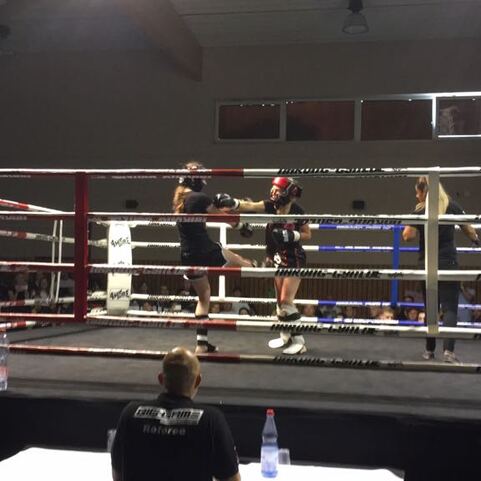 Zwei K&auml;mpfer im Boxring w&auml;hrend eines Wettkampfs, Zuschauer am Rand beobachten das Geschehen.