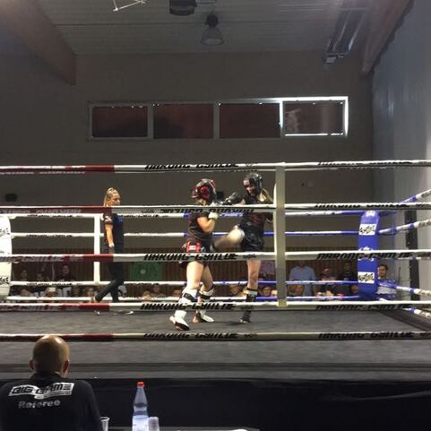 Zwei Boxer im Ring w&auml;hrend eines Wettkampfs, Zuschauer im Hintergrund, Trainer beobachtet.