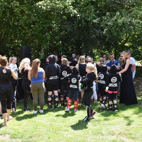 Gruppe von Personen in Sportkleidung versammelt im Freien, umgeben von B&auml;umen und Zuschauer im Hintergrund.