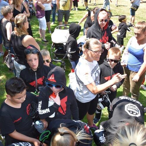 Gruppe von Kindern und Erwachsenen in sportlicher Kleidung mit Kapuzen auf einem Freiluftgel&auml;nde, umgeben von Zuschauern.