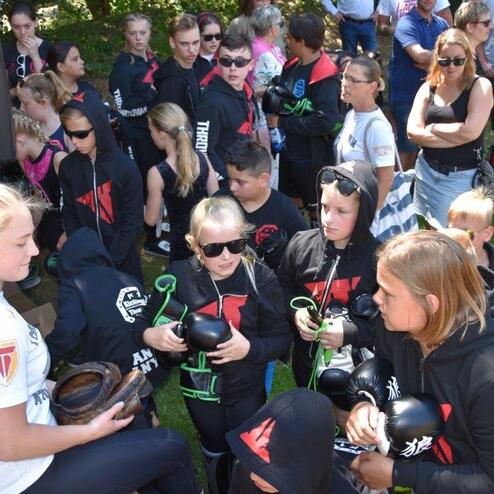 Gruppe von Kindern in Sportbekleidung versammelt sich um Trainerin, die ihnen Boxhandschuhe zeigt.