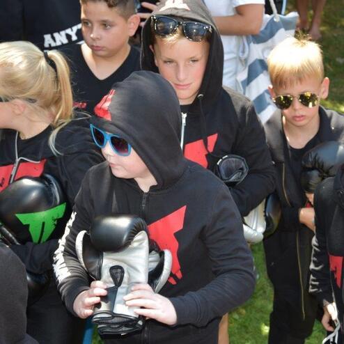 Gruppe von Kindern in schwarzen Hoodie-Jacken mit Boxhandschuhen, teils mit Sonnenbrillen, stehen auf einer Wiese.