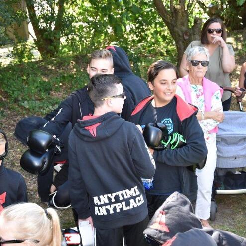 Gruppe von Jugendlichen in Sportkleidung mit Boxhandschuhen, umgeben von Zuschauern in einem gr&uuml;nen Park.