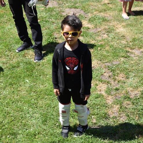 Junge mit Sonnenbrille und Spider-Man-Shirt steht auf einer gr&uuml;nen Wiese, umgeben von anderen Personen.