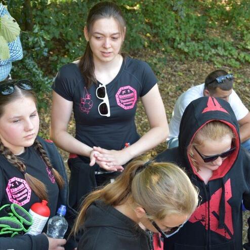 F&uuml;nf Jugendliche stehen im Freien, zwei sind mit Kapuzenpullovern bekleidet und diskutieren aufmerksam.