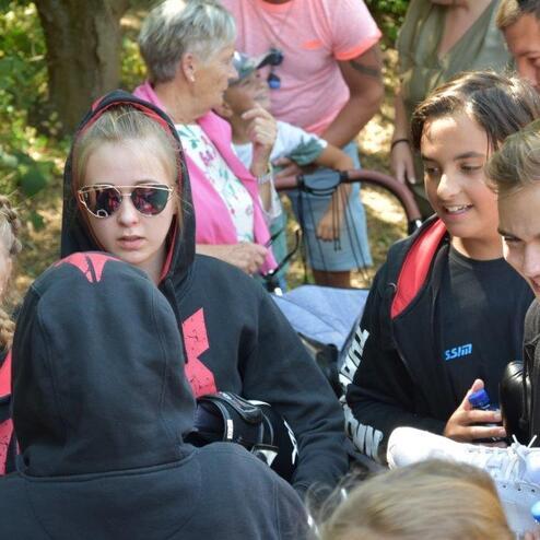 Gruppe junger Leute in Sportbekleidung diskutiert unter B&auml;umen, w&auml;hrend Erwachsene im Hintergrund sitzen.