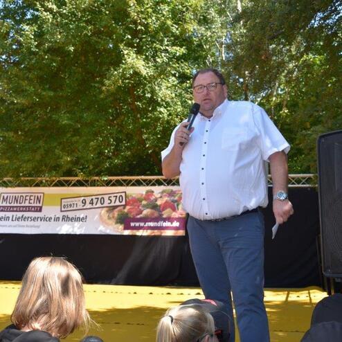 Mann spricht mit Mikrofon auf einer B&uuml;hne vor einem Publikum, umgeben von B&auml;umen und einem Banner.