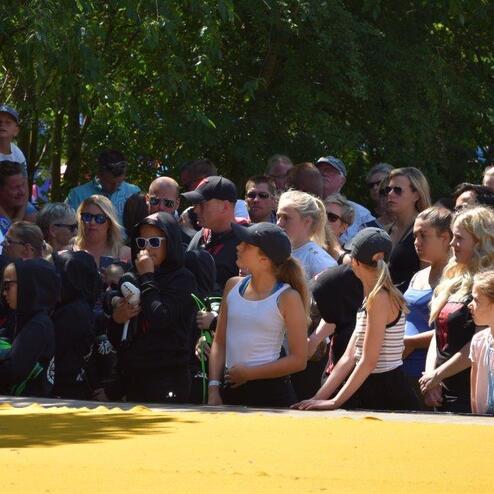 Wohlbeleuchtete Menschenmenge schaut gebannt auf eine Veranstaltung im Freien, umgeben von B&auml;umen und einer gelben Fl&auml;che.