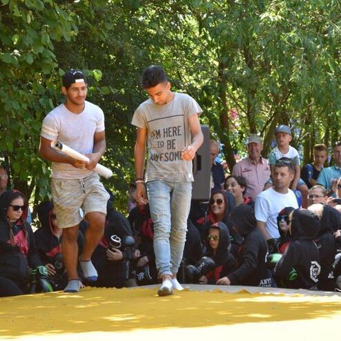 Zwei Jugendliche gehen auf einer gelben B&uuml;hne, umgeben von einem Publikum im Freien. B&auml;ume und Zuschauer sind im Hintergrund.