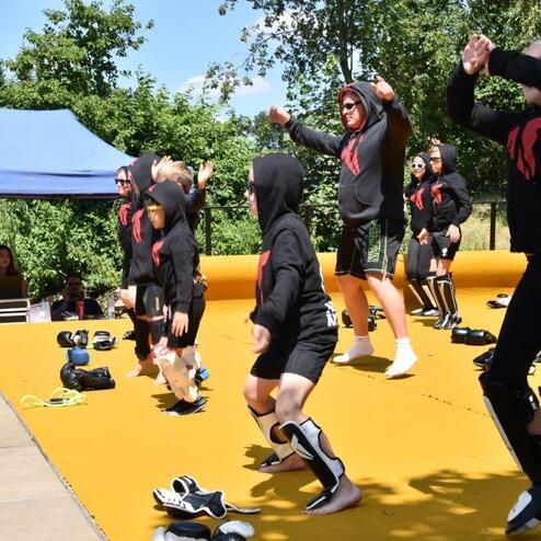 Kinder in schwarzen Kapuzenpullis tanzen auf einer gelben Fl&auml;che, umgeben von Zuschauern und B&auml;umen.