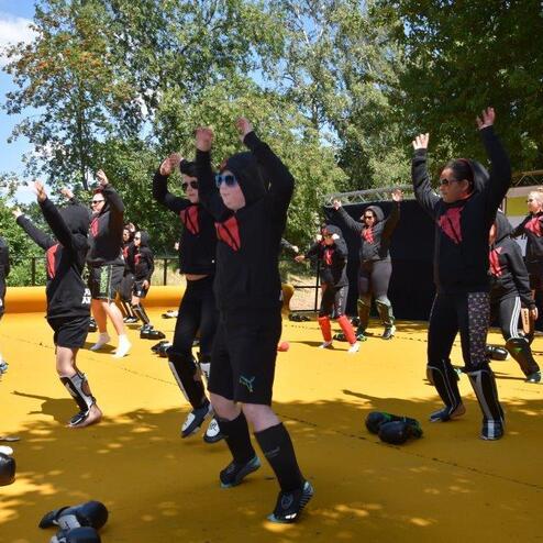 Gruppe von Menschen in Sportkleidung f&uuml;hrt Trainings&uuml;bungen auf einer gelben Fl&auml;che unter blauem Himmel aus.