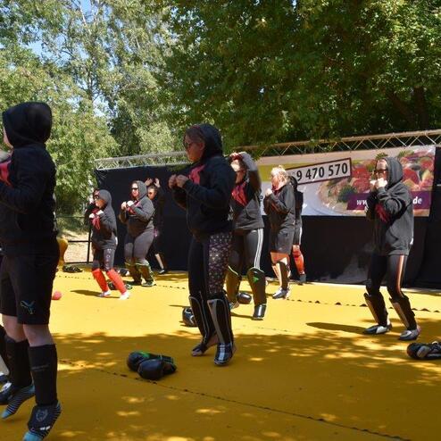 Gruppentraining mit mehreren Personen in Sportkleidung auf einer gelben Fl&auml;che, umgeben von B&auml;umen und einer Wand mit Plakat.