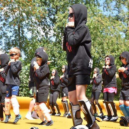 Gruppe von Kindern in Sportkleidung und Kapuzenpullis, die auf einer gelben Fl&auml;che &Uuml;bungen ausf&uuml;hren.