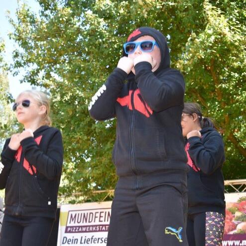 Tanzende Kinder in schwarzen Sweatshirts und Sonnenbrillen bei einer Veranstaltung im Freien.