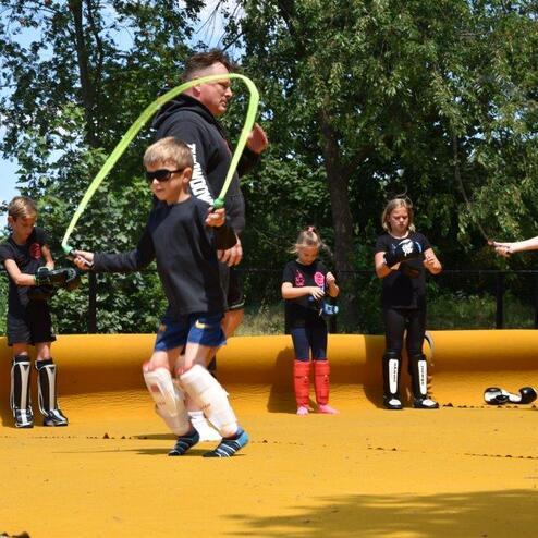 Kinder springen Seil auf einem gelben Trainingsbelag, w&auml;hrend ein Trainer Anweisungen gibt. B&auml;ume und Himmel im Hintergrund.