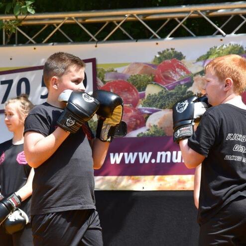 Zwei Kinder im Boxsack stehen sich gegen&uuml;ber und tragen Boxhandschuhe, im Hintergrund eine Werbebanner.