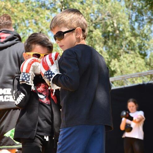 Zwei Kinder in Boxhandschuhen mit Sonnenbrillen stehen nah beieinander, w&auml;hrend Erwachsene im Hintergrund trainieren.