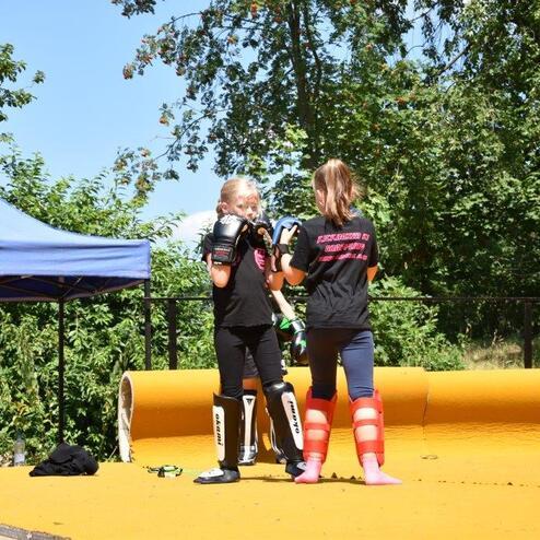Zwei M&auml;dchen in Trainingskleidung &uuml;ben mit Boxhandschuhen auf einer gelben Sportfl&auml;che im Freien.