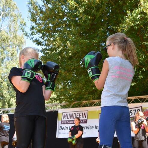 Zwei M&auml;dchen in Boxhandschuhen stehen sich im Freien, umgeben von weiteren Teilnehmenden bei einer Boxveranstaltung.