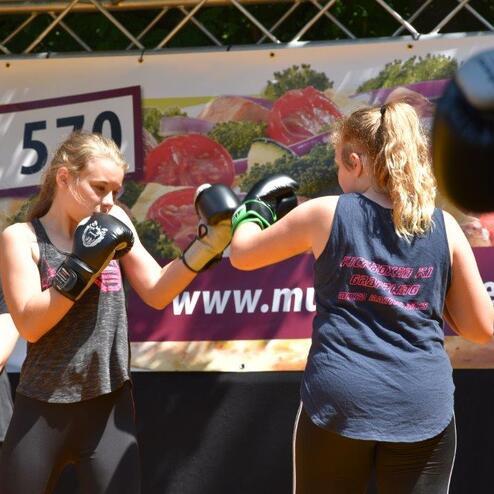 Zwei M&auml;dchen im Boxtraining tauschen Schl&auml;ge aus, w&auml;hrend ein Plakat im Hintergrund gut sichtbar ist.