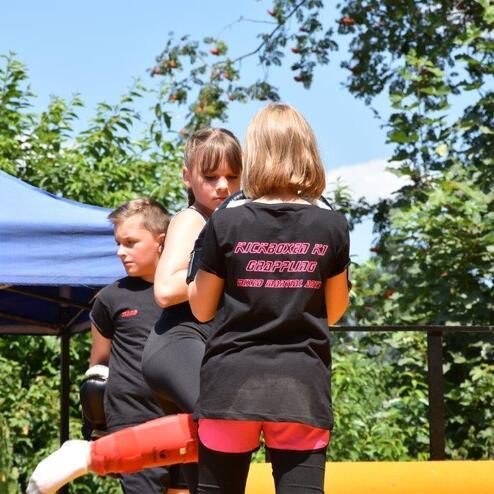 Zwei M&auml;dchen in Sportkleidung &uuml;ben Kampfsporttechniken in einer sonnigen Umgebung mit gr&uuml;nen B&auml;umen im Hintergrund.