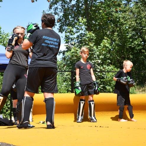 Gruppentraining von Kindern im Kampfsport auf einer gelben Matte, w&auml;hrend ein Trainer in Schwarz technische &Uuml;bungen zeigt.