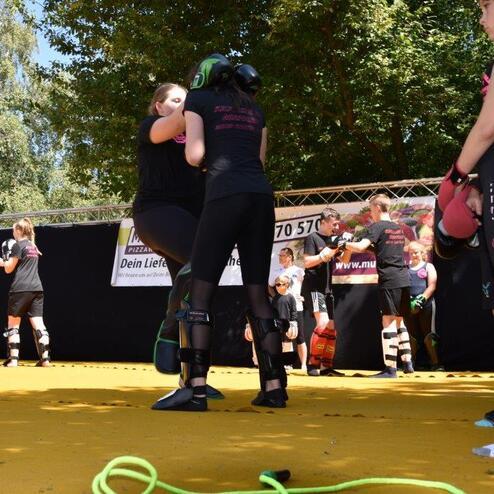 Zwei Frauen im Kampfsportanzug &uuml;ben eine Technik auf einer gelben Trainingsfl&auml;che, umgeben von weiteren Sportlern.