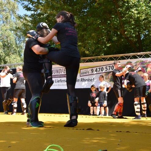 Zwei Personen im Kampfsporttraining auf einer gelben Matte, umgeben von anderen Trainierenden unter einem Werbebanner.