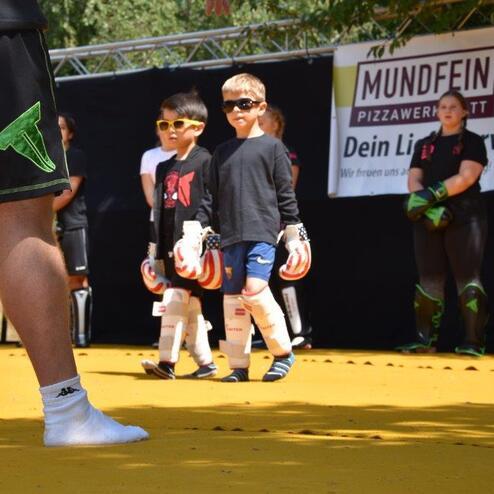 Zwei Kinder mit Torwarthandschuhen und Sonnenbrillen stehen auf einem gelben Sportplatz, bereit f&uuml;r das Training.