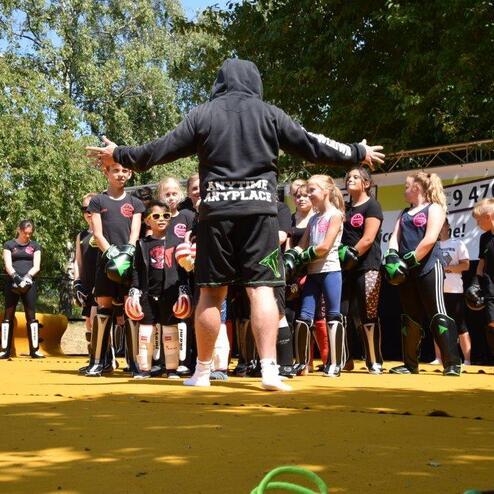 Trainer spricht mit einer Gruppe von Kindern auf einer Sportfl&auml;che, umgeben von nat&uuml;rlicher Umgebung.