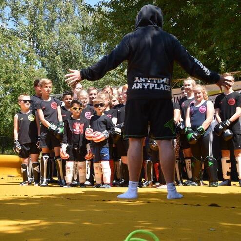 Gruppe von Kindern in Sportkleidung h&ouml;rt einem Trainer auf einer gelben Trainingsfl&auml;che aufmerksam zu.