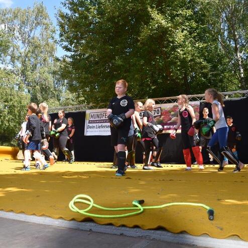Kinder in Sportkleidung trainieren auf einer gelben Fl&auml;che im Freien, umgeben von B&auml;umen und Zuschauern.