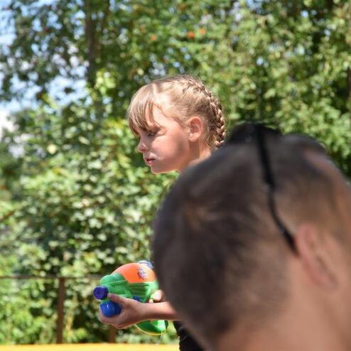 M&auml;dchen mit geflochtenem Haar h&auml;lt eine Wasserpistole, im Hintergrund unscharf ein Erwachsener und B&auml;ume.