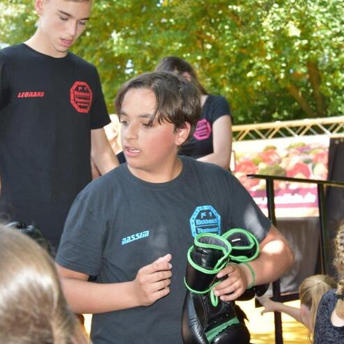 Jugendlicher in schwarzem T-Shirt h&auml;lt Boxhandschuhe, umgeben von anderen Kindern in einem Freiluftumfeld.