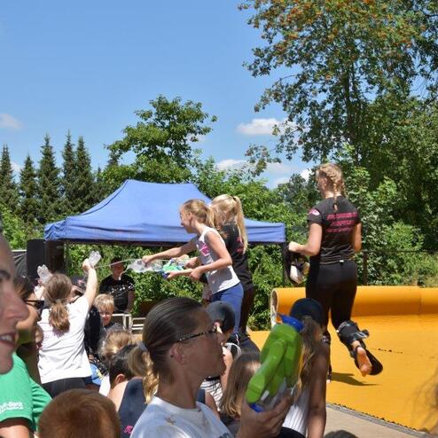 Gruppenevent im Freien: Kinder und Jugendliche stehen um einen blauen Pavillon, mit einer H&uuml;pfburg im Hintergrund.