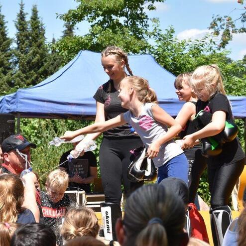 Vier M&auml;dchen in Sportoutfits stehen auf einer B&uuml;hne, umgeben von Zuschauern, w&auml;hrend eines Outdoor-Events im Freien.