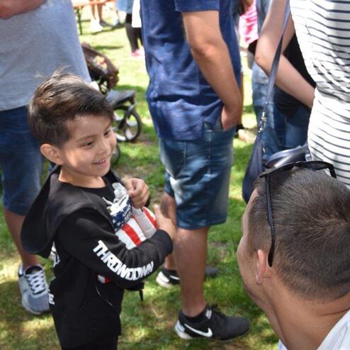 Ein l&auml;chelndes Kind steht im Gespr&auml;ch mit einem Erwachsenen, umgeben von einer Menschenmenge auf einer Wiese.