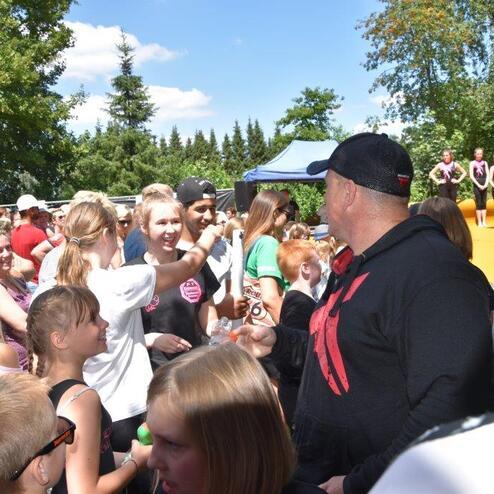 Menschenmenge bei einem Outdoor-Event, einige Kinder und Erwachsene interagieren im Vordergrund, gr&uuml;ne B&auml;ume im Hintergrund.
